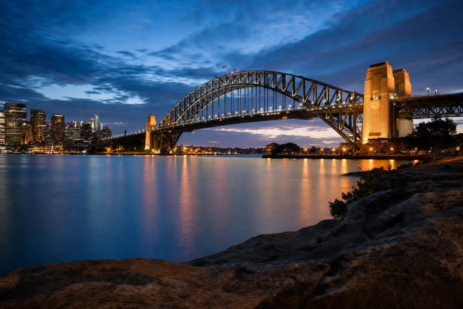 Sydney Harbour Bridge at blue hour seen from Mrs Macquarie's Chair across calm harbour water